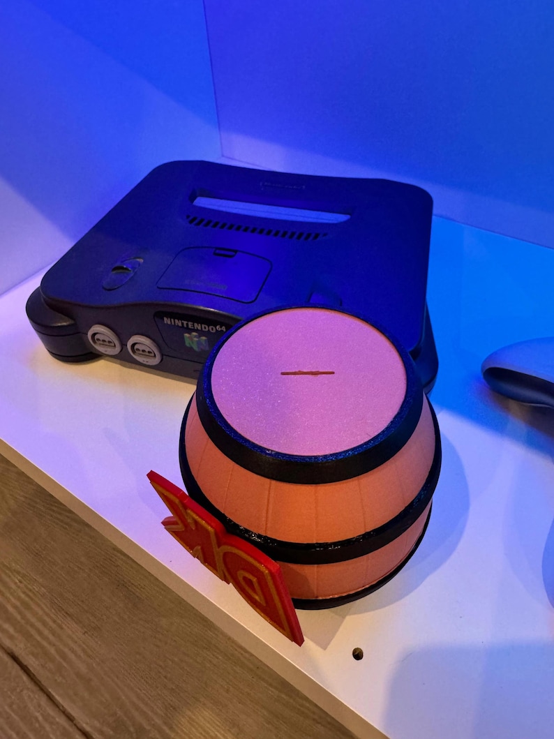 May include: A black Nintendo 64 game console with a barrel-shaped object and a red and gold DK logo. The barrel is orange with black bands. The DK logo is red with gold accents. The console is on a white surface.