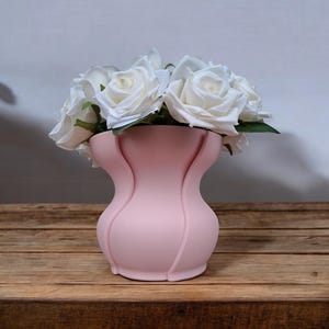 Puede incluir: Un jarr&oacute;n rosa p&aacute;lido con un dise&ntilde;o curvo &uacute;nico, que contiene un ramo de rosas blancas. El jarr&oacute;n tiene una superficie texturizada y est&aacute; sobre una mesa de madera. Las rosas tienen hojas verdes.