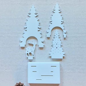 May include: A white Christmas-themed decorative set featuring three stylized fir trees, a reindeer silhouette, and a rectangular base with horizontal cutouts. A pine cone is also present. The pieces are arranged on a light-colored surface.