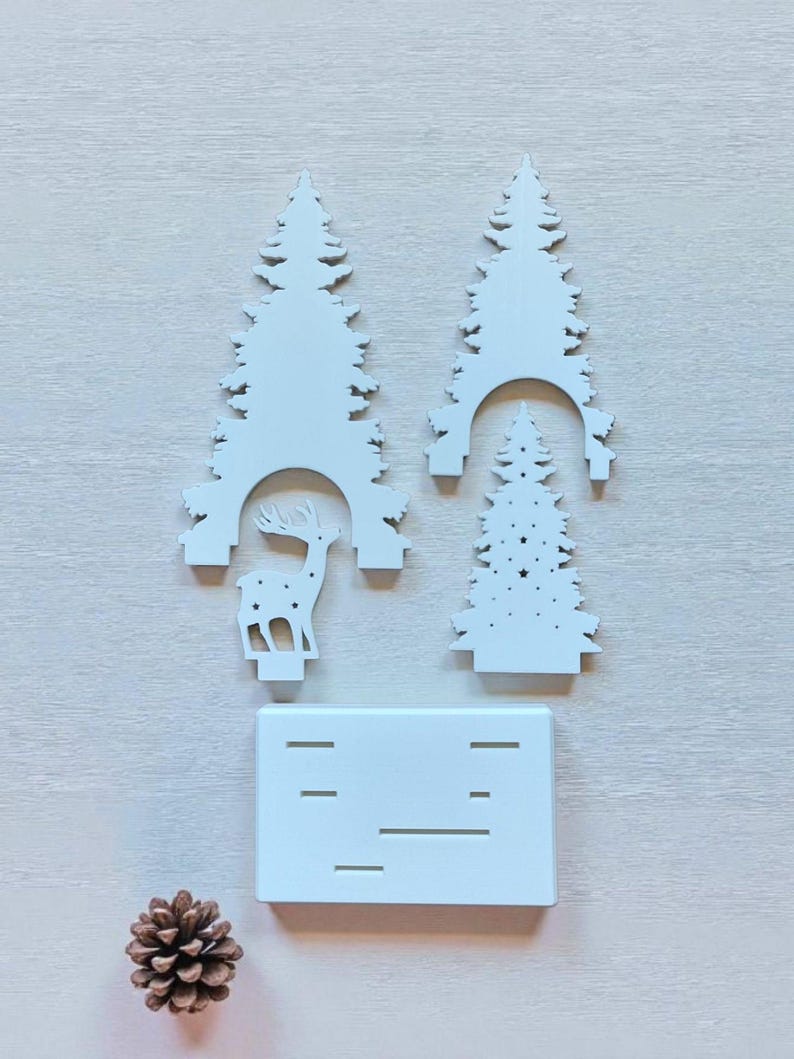 May include: A white Christmas-themed decorative set featuring three stylized fir trees, a reindeer silhouette, and a rectangular base with horizontal cutouts. A pine cone is also present. The pieces are arranged on a light-colored surface.
