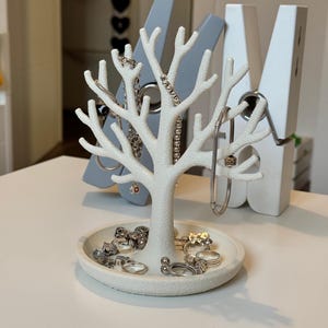 May include: A white jewelry tree with a round base and branches, displaying rings and necklaces. The tree is surrounded by three large, decorative clothespins in gray and white. The jewelry is silver-toned.