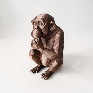 May include: A bronze-colored, geometric sculpture of a monkey in a seated position with hands clasped. The monkey has a thoughtful expression and is set against a white background. The sculpture's design features many flat planes.
