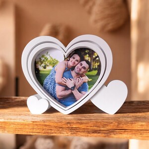 May include: A white heart-shaped photo frame with a picture of a couple embracing. The frame has a layered design and two small heart-shaped supports. The photo shows a couple smiling and hugging each other.
