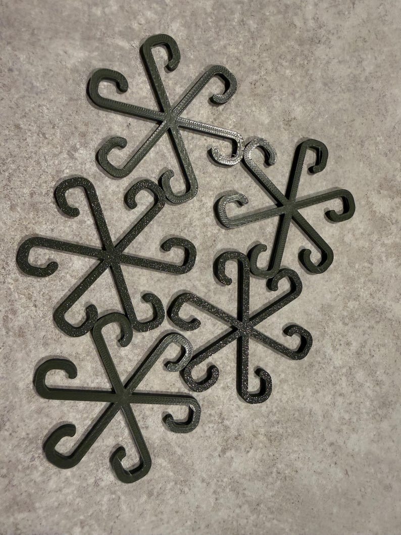 May include: A collection of six dark green, snowflake-shaped decorative items. Each piece features six curved arms extending from a central point, with a looped design at the end of each arm. The items are arranged on a light gray surface.