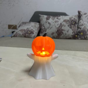 May include: A Halloween-themed decoration featuring a glowing orange jack-o'-lantern head atop a white ghost figure. The pumpkin has carved-out eyes, nose, and mouth, and the ghost has outstretched arms. The light inside the pumpkin illuminates the details.