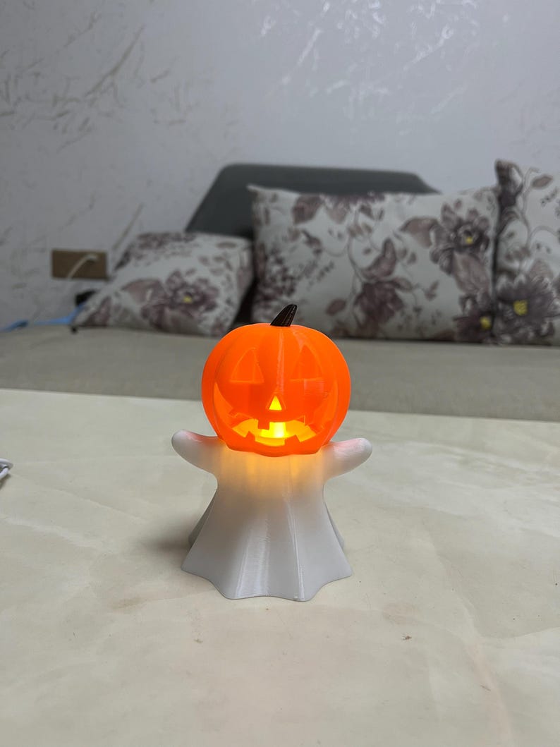 May include: A Halloween-themed decoration featuring a glowing orange jack-o'-lantern head atop a white ghost figure. The pumpkin has carved-out eyes, nose, and mouth, and the ghost has outstretched arms. The light inside the pumpkin illuminates the details.