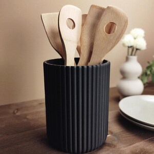 May include: A black, ribbed utensil holder filled with wooden spatulas and spoons. The kitchen tools are light brown and have rounded edges. The holder is on a wooden table, with a vase and plates in the background.