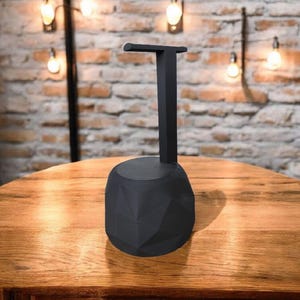 May include: A black headphone stand with a geometric base and a vertical support arm. The stand is made of a matte black material and is designed to hold headphones. The stand is on a wooden table.
