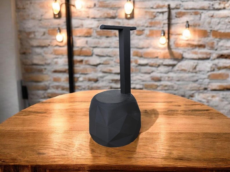 May include: A black headphone stand with a geometric base and a vertical support arm. The stand is made of a matte black material and is designed to hold headphones. The stand is on a wooden table.