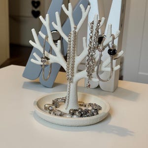 May include: A white tree-shaped jewelry organizer with a round base, holding various silver necklaces, bracelets, and rings. The organizer is set against a backdrop of large gray and white clothespins.