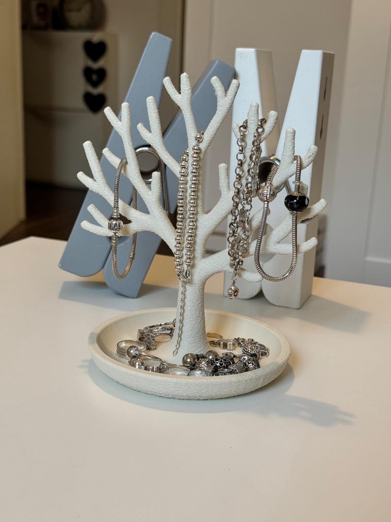 May include: A white tree-shaped jewelry organizer with a round base, holding various silver necklaces, bracelets, and rings. The organizer is set against a backdrop of large gray and white clothespins.