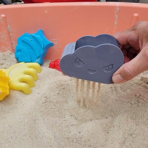 May include: A hand holding a gray cloud-shaped sand sifter, with sand falling through. Other colorful sand molds, including a blue fish, yellow hand, and red shape, are scattered in the sand. The scene is set in a pink plastic sandbox.
