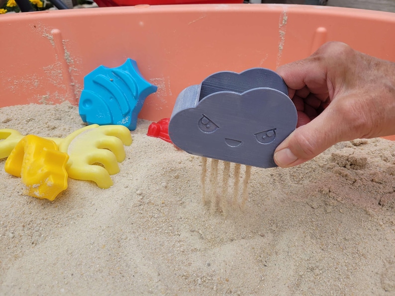 May include: A hand holding a gray cloud-shaped sand sifter, with sand falling through. Other colorful sand molds, including a blue fish, yellow hand, and red shape, are scattered in the sand. The scene is set in a pink plastic sandbox.