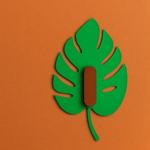 以下が含まれることがあります： オレンジ色の背景に、茶色のフックが付いた緑色のモンステラ葉の形をしたウォールフック。 フックは葉っぱに似せてデザインされており、どんな空間にも自然をイメージした装飾を添えます。