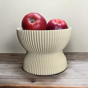 May include: A cream-colored, ribbed bowl holds two red apples. The bowl has a unique hourglass shape and sits on a wooden surface. The apples are a deep red color.