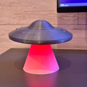 May include: A gray, 3D-printed UFO-shaped lamp with a red glowing base. The lamp has a classic flying saucer design with a domed top and small, round details around the rim. The base is a cone shape, emitting a bright red light.