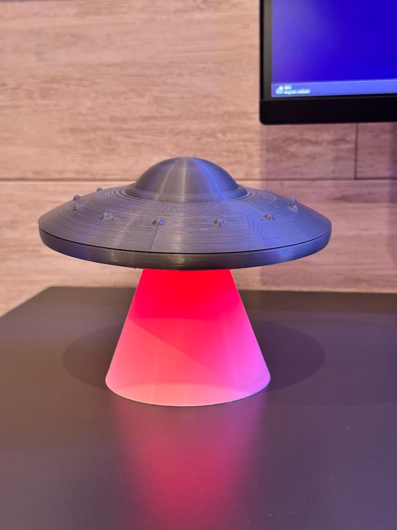 May include: A gray, 3D-printed UFO-shaped lamp with a red glowing base. The lamp has a classic flying saucer design with a domed top and small, round details around the rim. The base is a cone shape, emitting a bright red light.