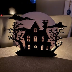 Pu&ograve; includere: Silhouette nera di una casa infestata con finestre illuminate, ambientata su una luna pallida e nuvole scure. Due alberi spogli incorniciano la casa, creando una spettrale decorazione di Halloween. La casa ha un camino e un tetto dettagliato.