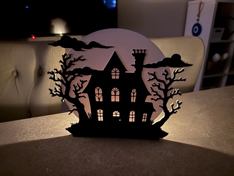 Pu&ograve; includere: Silhouette nera di una casa infestata con finestre illuminate, ambientata su una luna pallida e nuvole scure. Due alberi spogli incorniciano la casa, creando una spettrale decorazione di Halloween. La casa ha un camino e un tetto dettagliato.