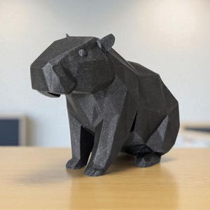 May include: A black, geometric capybara sculpture. The figure is made of many flat, angled surfaces, giving it a faceted appearance. The capybara is sitting on a light-colored wooden surface.