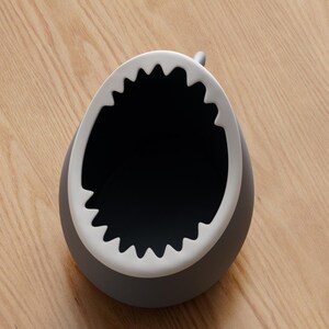 May include: A gray, shark-shaped container with a white, jagged rim resembling teeth. The interior is black. The container sits on a light brown wooden surface.