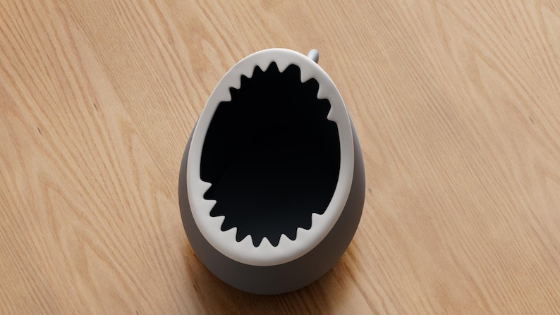 May include: A gray, shark-shaped container with a white, jagged rim resembling teeth. The interior is black. The container sits on a light brown wooden surface.