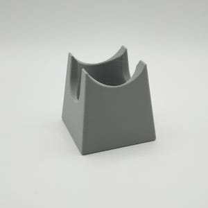 May include: A gray, 3D-printed stand with a square base and two curved supports. The stand is designed to hold an item, with a hollowed-out space between the supports. The object is made of a solid, matte material.