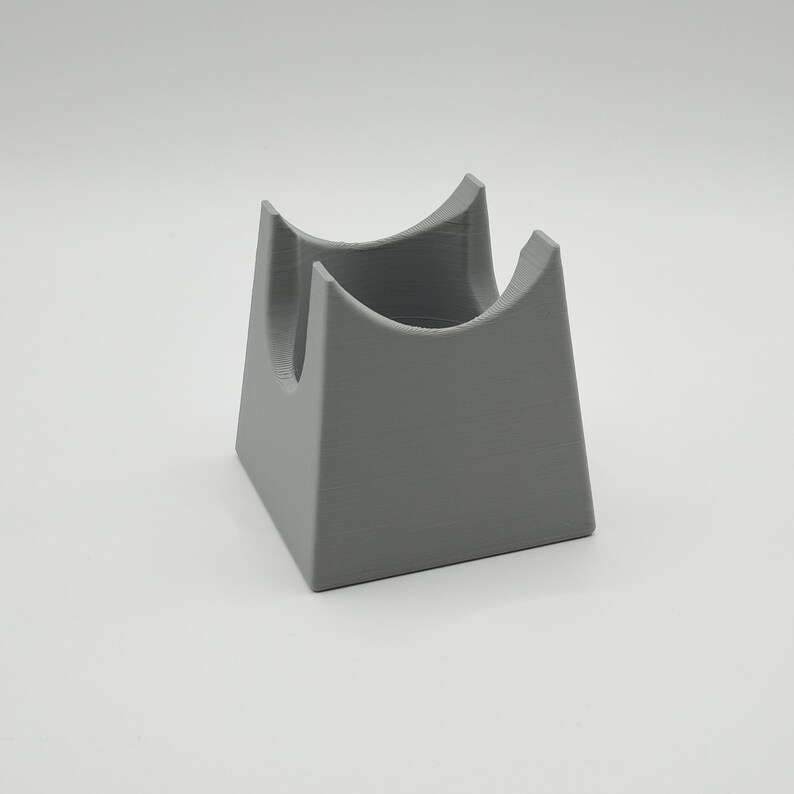 May include: A gray, 3D-printed stand with a square base and two curved supports. The stand is designed to hold an item, with a hollowed-out space between the supports. The object is made of a solid, matte material.