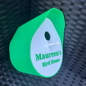 May include: A green and white birdhouse with a rounded roof and a small hole for birds to enter. The birdhouse has the text "Maureen's Bird House" printed on the front.