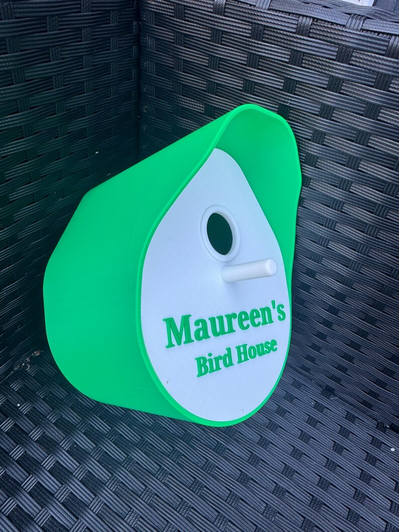 May include: A green and white birdhouse with a rounded roof and a small hole for birds to enter. The birdhouse has the text "Maureen's Bird House" printed on the front.