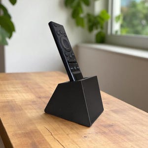 May include: A black remote control rests in a black, geometric stand on a wooden surface. The remote has multiple buttons and a circular control pad. The stand is a simple, modern design, with a triangular base.