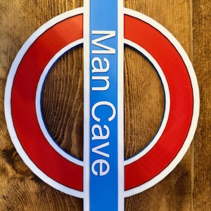 May include: A sign with a red circle, white border, and blue vertical rectangle. The words "Man Cave" are printed vertically in white on the blue rectangle. The sign is mounted on a wooden surface.