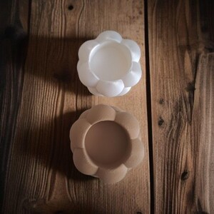 May include: Two flower-shaped candle holders. One is white, the other is beige. The holders are made of a smooth material and sit on a wooden surface. The candle holders have a round center and a petal-like design around the edges.