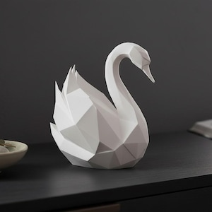 May include: A white geometric swan sculpture with a modern, angular design. The swan is positioned on a dark surface, with a shallow bowl containing stones and greenery in the background. A book is also visible.