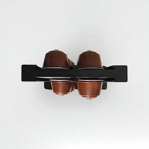 May include: A black metal coffee pod holder containing six brown coffee pods. The holder is rectangular with individual slots. The pods are a dark brown colour, likely filled with coffee. The background is plain white.