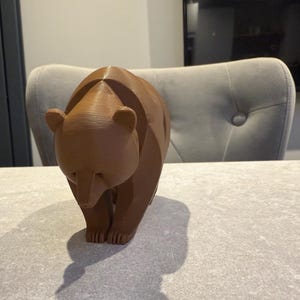 May include: A brown bear figurine with a geometric design. The bear is standing on all fours, with its head slightly lowered. The figurine is made of a solid material and has a smooth finish. The bear is a decorative item.