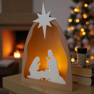 May include: A wooden nativity scene with a white star. The scene shows the silhouettes of Mary, Joseph, and baby Jesus, lit by a warm glow. A Christmas tree with soft lights is in the background.