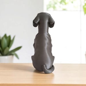 May include: A dark gray dog figurine, viewed from the back, sits on a light brown wooden surface. The dog has large, floppy ears and a curled tail. The background is blurred, with a glimpse of a window and greenery.
