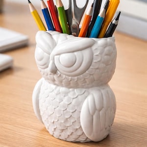 May include: A white ceramic owl-shaped desk organizer filled with colorful pencils, pens, and scissors. The owl has a detailed, textured surface and is sitting on a wooden desk. A decorative and functional item for office or home.