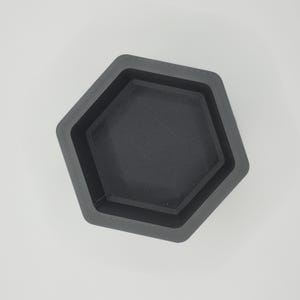 May include: A dark gray, hexagonal container with a textured interior. The container has a stepped design, with a smaller hexagon nested inside the larger one. The object is made of a matte material and sits on a white surface.