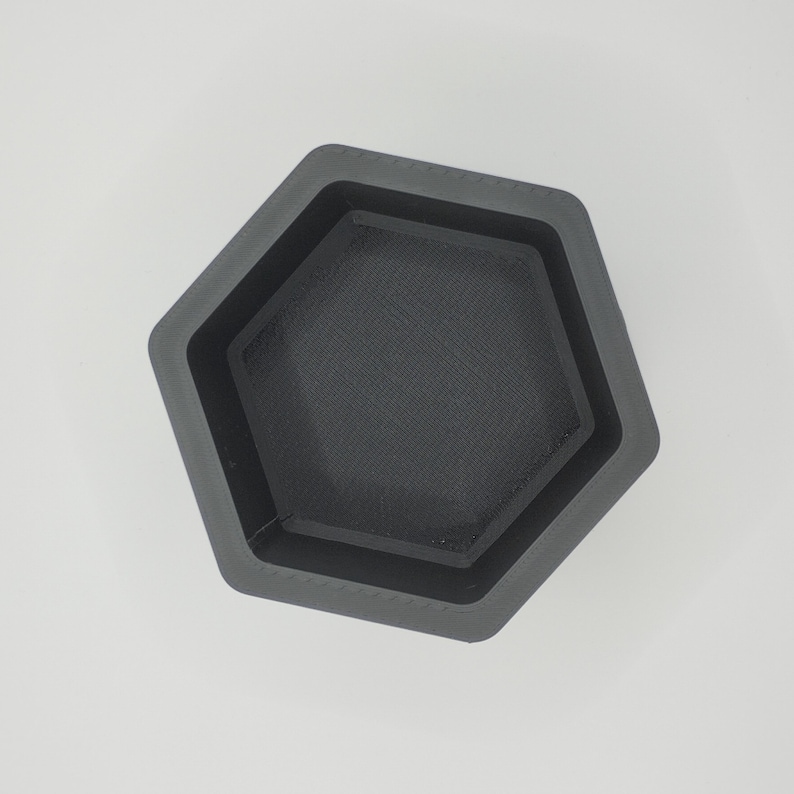 May include: A dark gray, hexagonal container with a textured interior. The container has a stepped design, with a smaller hexagon nested inside the larger one. The object is made of a matte material and sits on a white surface.