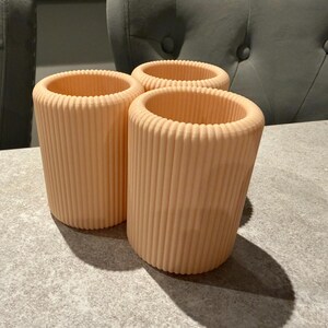 May include: Three peach-colored, cylindrical containers with vertical ribbed detailing. The containers are open at the top and sit on a gray surface. The containers are likely for holding items.