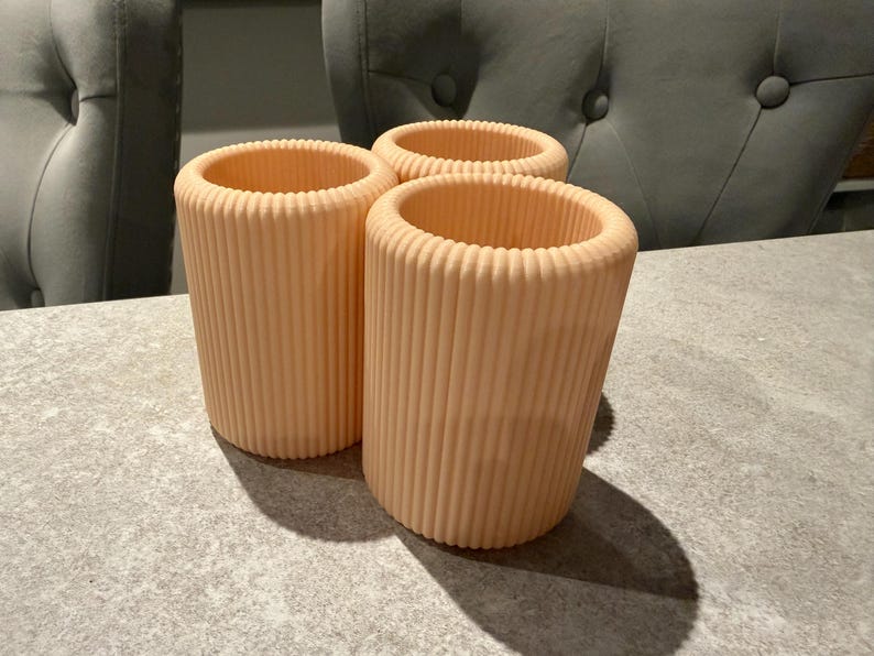 May include: Three peach-colored, cylindrical containers with vertical ribbed detailing. The containers are open at the top and sit on a gray surface. The containers are likely for holding items.