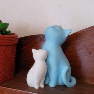 May include: Two cat figurines, one white and one light blue, sit side-by-side on a wooden shelf. A potted succulent plant in a terracotta pot is to the left. The cat figurines are smooth and stylized.