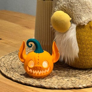 May include: An orange, 3D-printed jack-o'-lantern with a teal stem and large, pointed ears. The pumpkin has a wide, toothy grin and glowing eyes. It is sitting on a woven mat, with a decorative vase and gnome in the background.