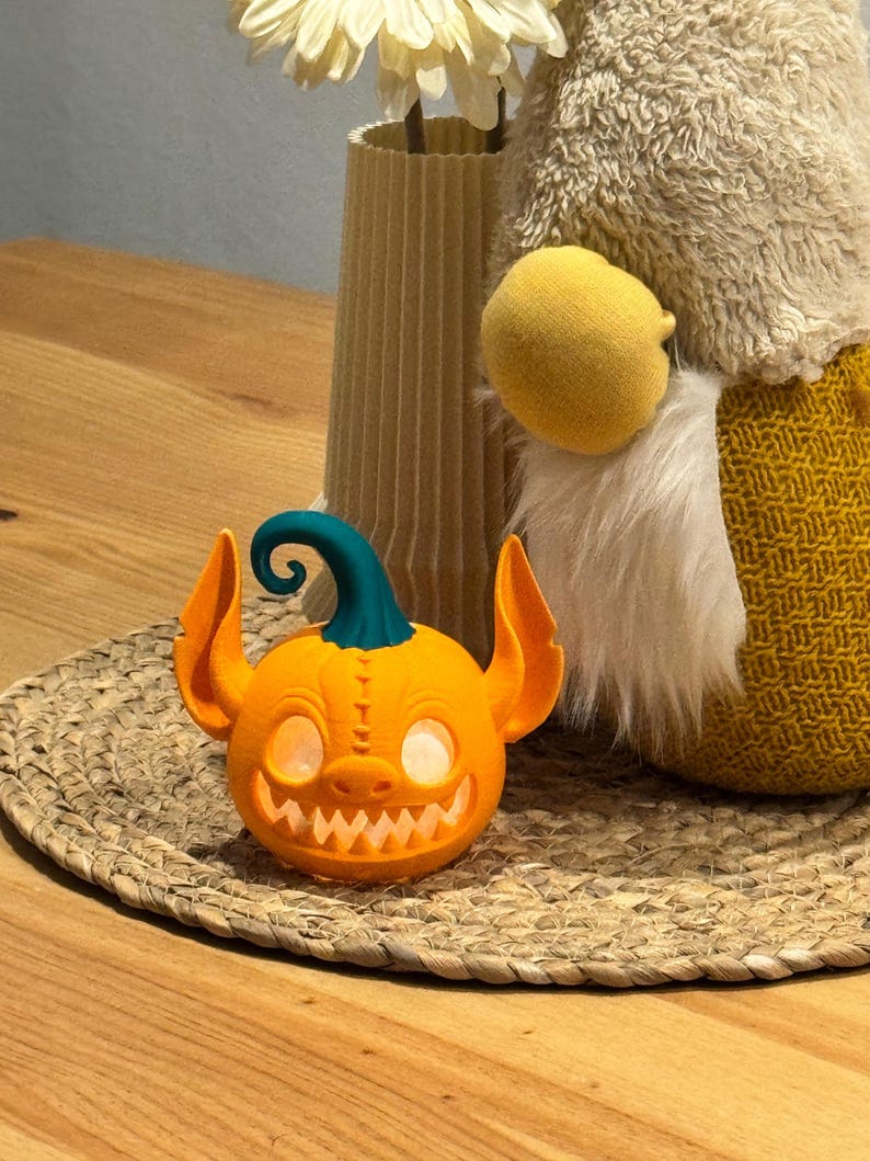 May include: An orange, 3D-printed jack-o'-lantern with a teal stem and large, pointed ears. The pumpkin has a wide, toothy grin and glowing eyes. It is sitting on a woven mat, with a decorative vase and gnome in the background.