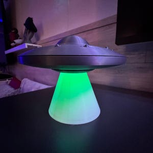 May include: A gray, 3D-printed UFO lamp with a green light emanating from the base. The lamp is sitting on a dark surface. The lamp has a classic flying saucer design.