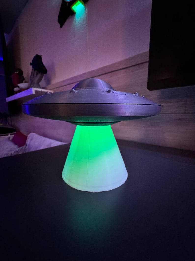 May include: A gray, 3D-printed UFO lamp with a green light emanating from the base. The lamp is sitting on a dark surface. The lamp has a classic flying saucer design.