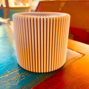 May include: A beige cylindrical planter with vertical ribbed detailing. The planter is made of a light-coloured material and sits on a wooden surface. The design features evenly spaced vertical lines, creating a textured appearance. The overall aesthetic is modern and minimalist.