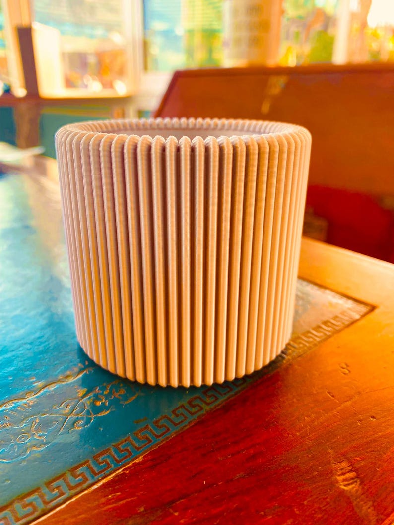 May include: A beige cylindrical planter with vertical ribbed detailing. The planter is made of a light-coloured material and sits on a wooden surface. The design features evenly spaced vertical lines, creating a textured appearance. The overall aesthetic is modern and minimalist.
